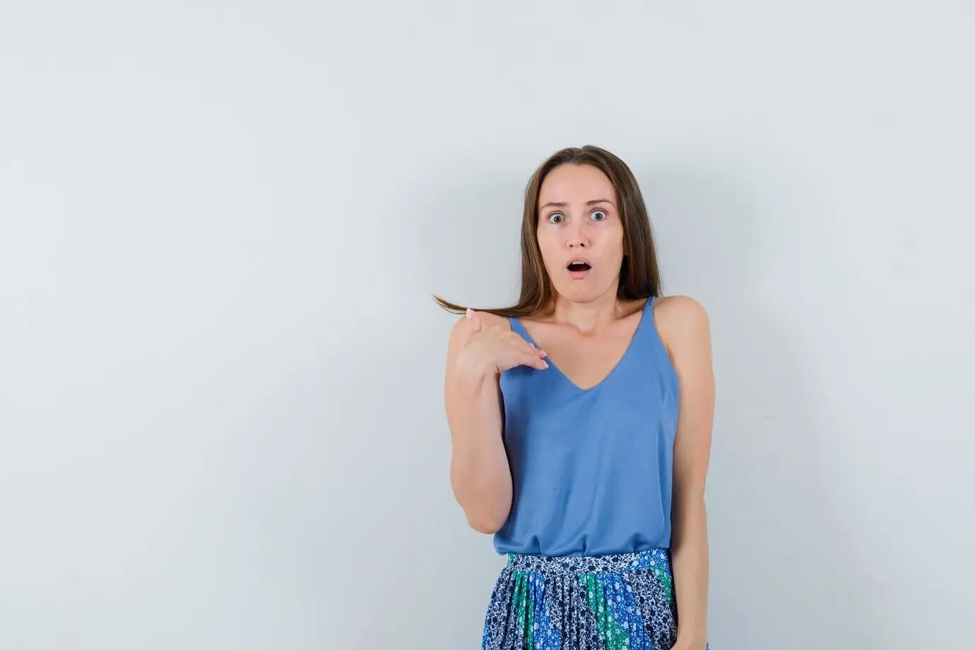 Молодая девушка указывает на себя, глядя в камеру с отвисшей челюстью; на ней блузка и юбка, выглядит нервной. Вид спереди.