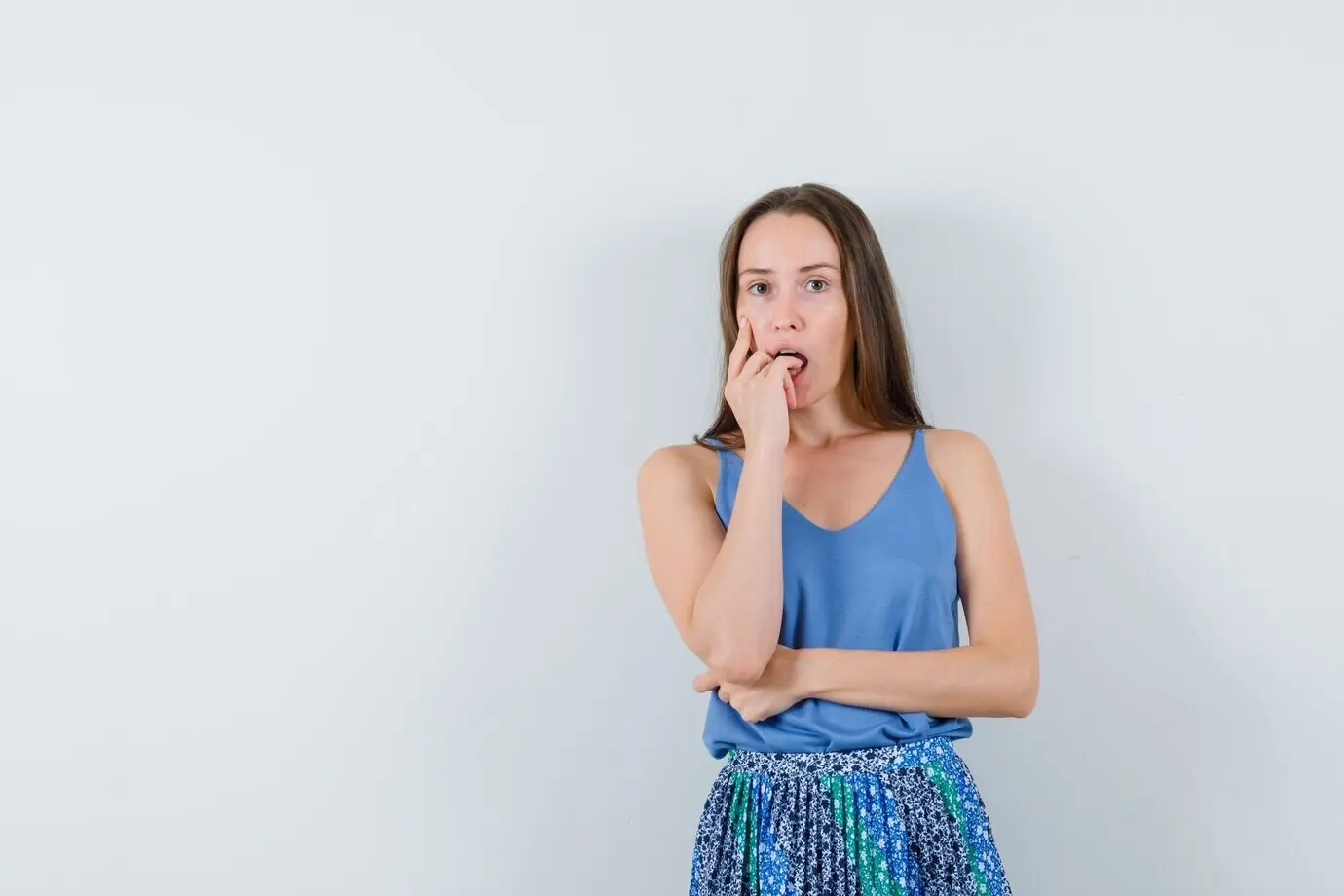 Молодая женщина в синей блузке и юбке прикусывает палец и задумчиво смотрит, вид спереди.
